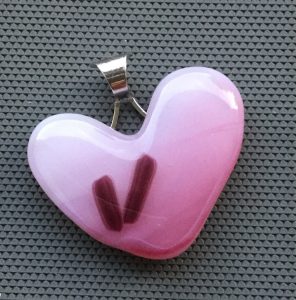 Ljusrosa hänge i hjärtform med 2 små lila streck. Silverfärgat bakstycke i form av hjärta